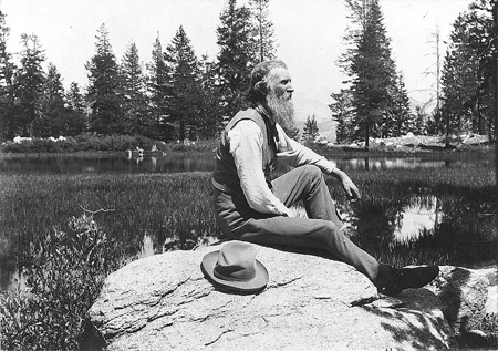 John Muir, full-length portrait, facing right, seated on rock with lake and trees in background